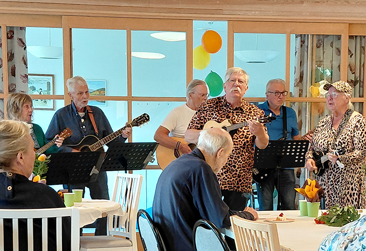 En grupp personer spelar musikaliskt instrument med en notställ. Flera ballonger i gul och grön färg syns.