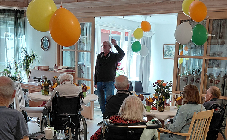 En man står och talar för en grupp människor sittandes runt bord med ballonger. Vissa personer sitter i rullstolar.