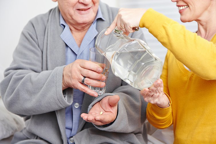 Äldre man tar sin medicin. Får hjälp med att hälla vatten i ett gals.